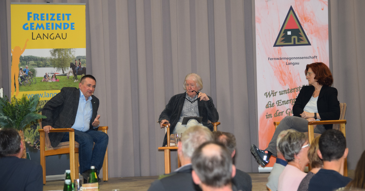 Drei Personen sitzen auf einer Bühne in Holzstühlen; Banner Freizeit Gemeinde Langau links, Fernwärmegenossenschaft Langau rechts.