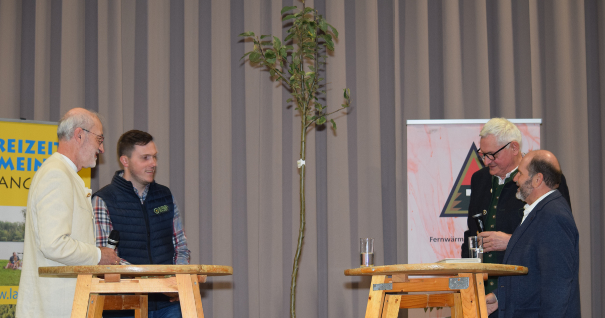 Vier Personen stehen an zwei hohen Holztischen auf einer Bühne; eine Person hält ein Mikrofon, zwei Gläser stehen bereit; Banner im Hintergrund.