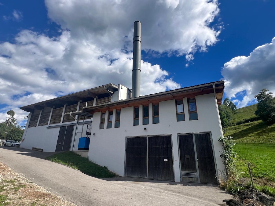 Industriegebäude mit hohem Schornstein, weißer Fassade, dunklen Toren, umliegender Grünfläche und blauem Himmel mit Wolken.