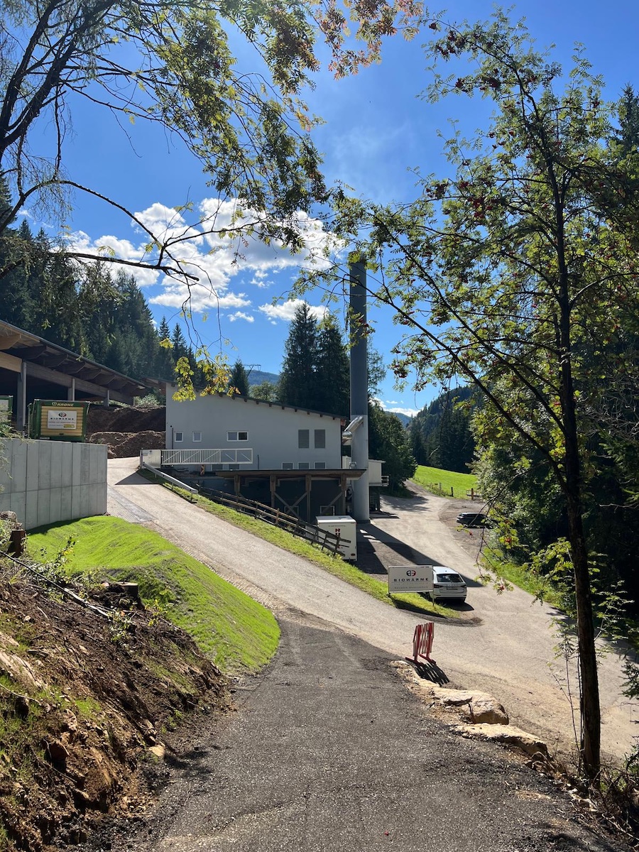 Industrieanlage mit hohem Schornstein, weißem Gebäude, Zufahrtsweg und Lieferwagen; umgeben von Bäumen, Hanglandschaft und blauem Himmel.