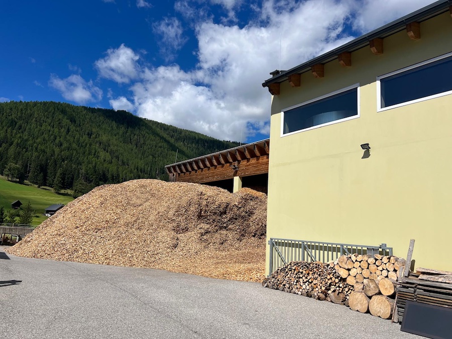 Großer Haufen Hackschnitzel neben gelbem Gebäude, davor Holzstapel, Metallzaun, asphaltierte Fläche und bewaldete Hügel unter blauem Himmel.