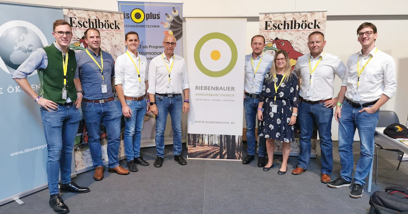 Neun Personen in Business-Casual vor Messestand mit Bannern; gelbe Lanyards, posieren für ein Foto.