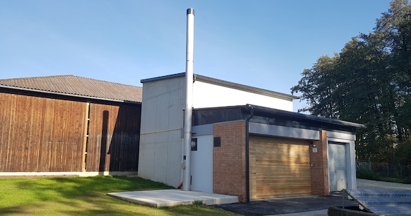 Kleines gemauertes Gebäude mit Rollladen-Tor, hohem Schornstein und weißem Anbau; links Holzbau, davor Grasfläche.