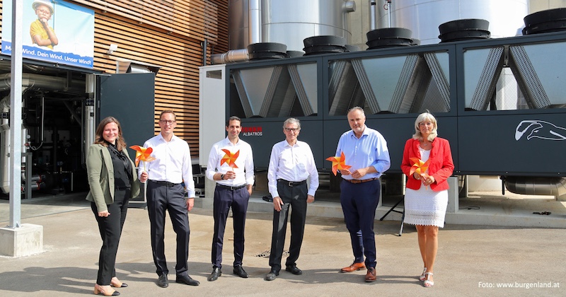 Sechs Personen stehen vor einer Industrieanlage und halten orange Pinwheels als Symbol für Windkraft.