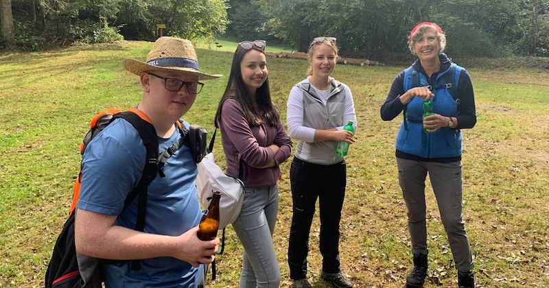Vier Personen beim Wandern im Freien; einer trägt Rucksack und Hut, zwei halten Trinkflaschen, weitere Person trägt Weste, fröhlich zusammen.