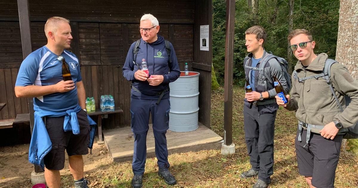 Vier Personen mit Rucksäcken stehen draußen vor einer Holzhütte, trinken Getränke aus Flaschen und führen ein Gespräch.