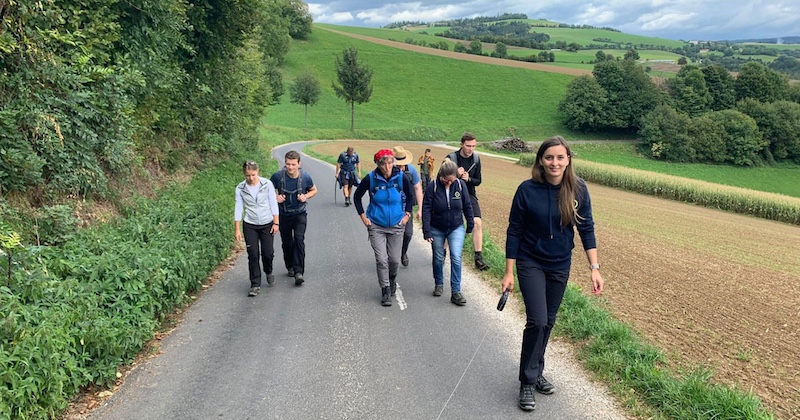 Gruppe von Personen läuft zu Fuß eine Landstraße entlang durch grüne Felder; im Hintergrund Hügel und Bäume.
