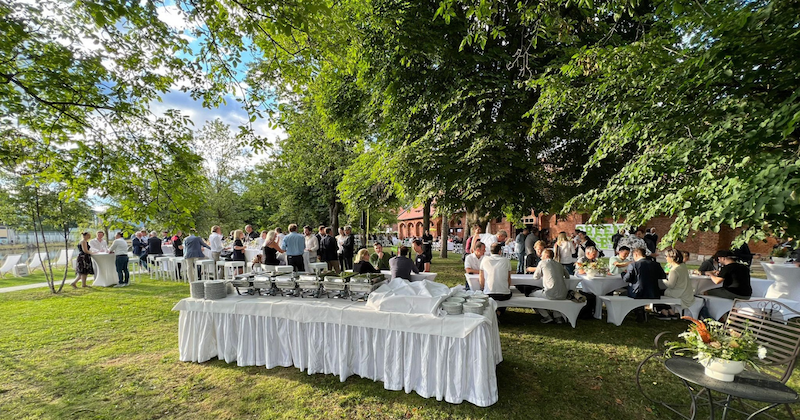 Outdoor-Gartenveranstaltung mit Buffet-Tisch, viele Gäste versammeln sich unter Bäumen.