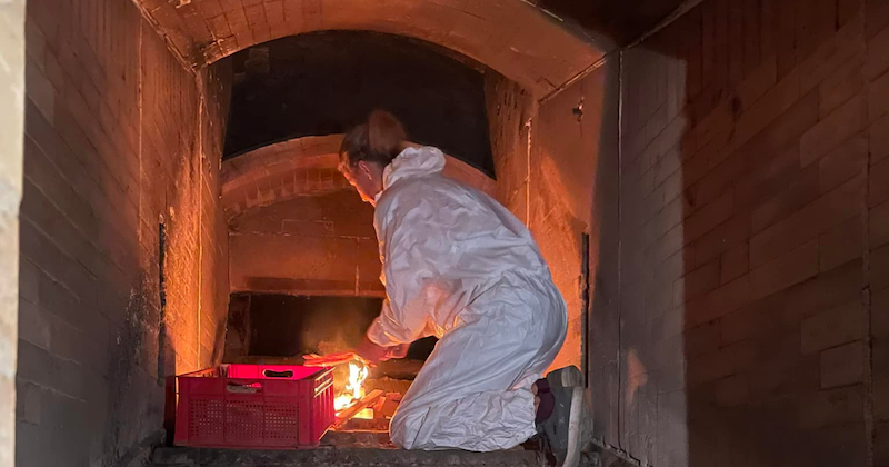 Person in weißem Schutzanzug kniet in einem Ziegel-Schacht vor brennender Flamme; rote Kiste daneben.