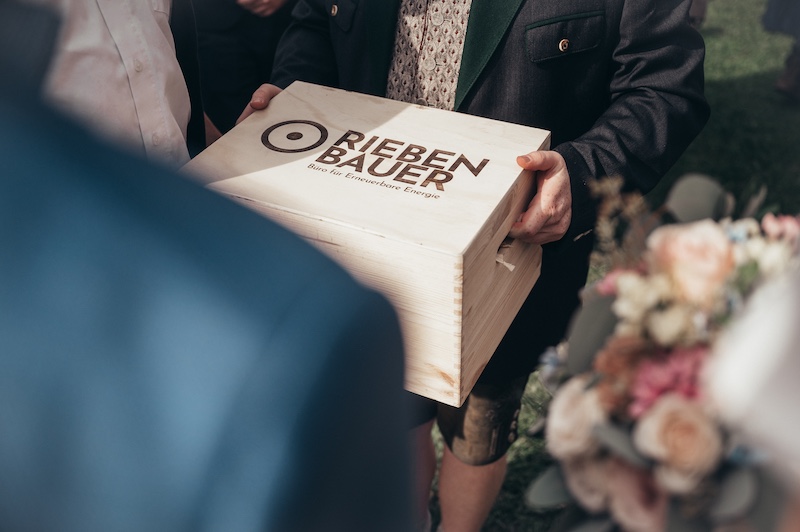 Holzkiste mit Logo 'RIEBENBAUER' und Unterzeile 'Büro für Erneuerbare Energie', von mehreren Personen getragen.