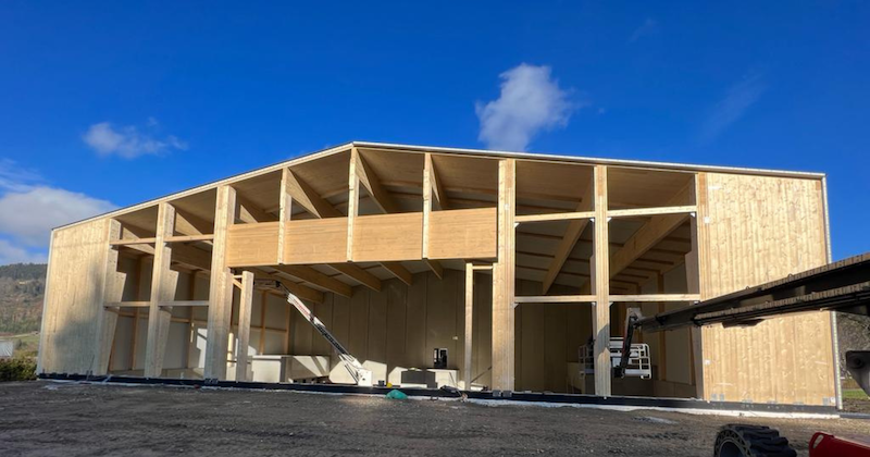 Holzrahmenbau im Rohbau mit offener Tragstruktur, Kran und Baumaschinen auf Baustelle, klarer blauer Himmel.