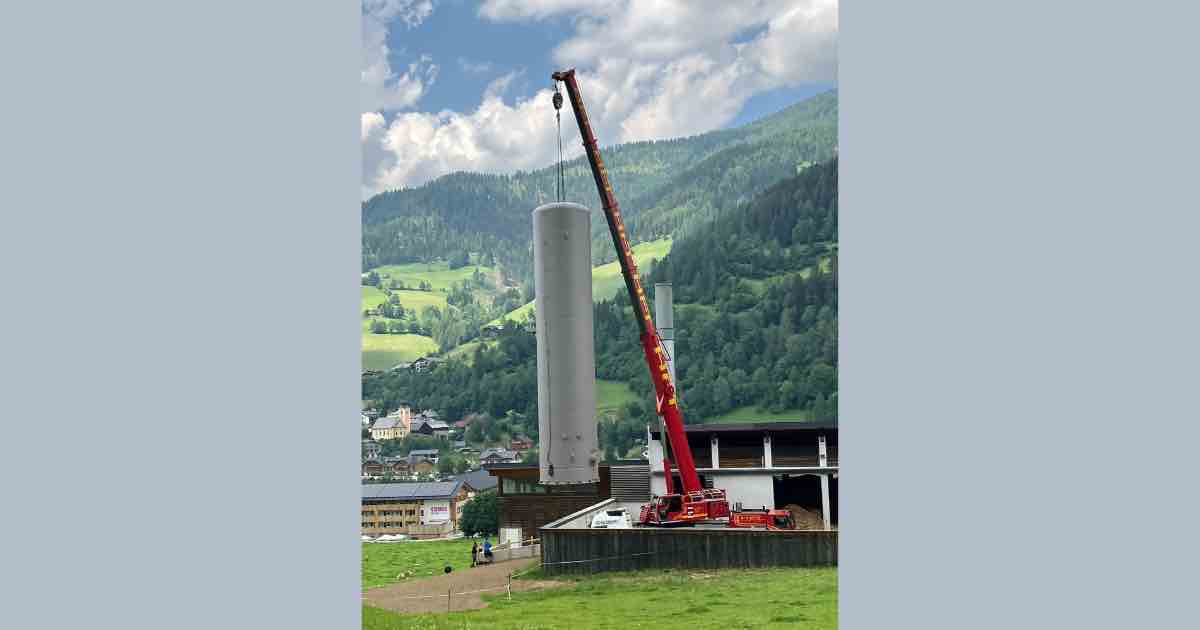 Kran hebt einen hohen grauen Pufferspeicher auf ein Bauwerk vor einer bergigen Landschaft mit Dorf im Hintergrund.