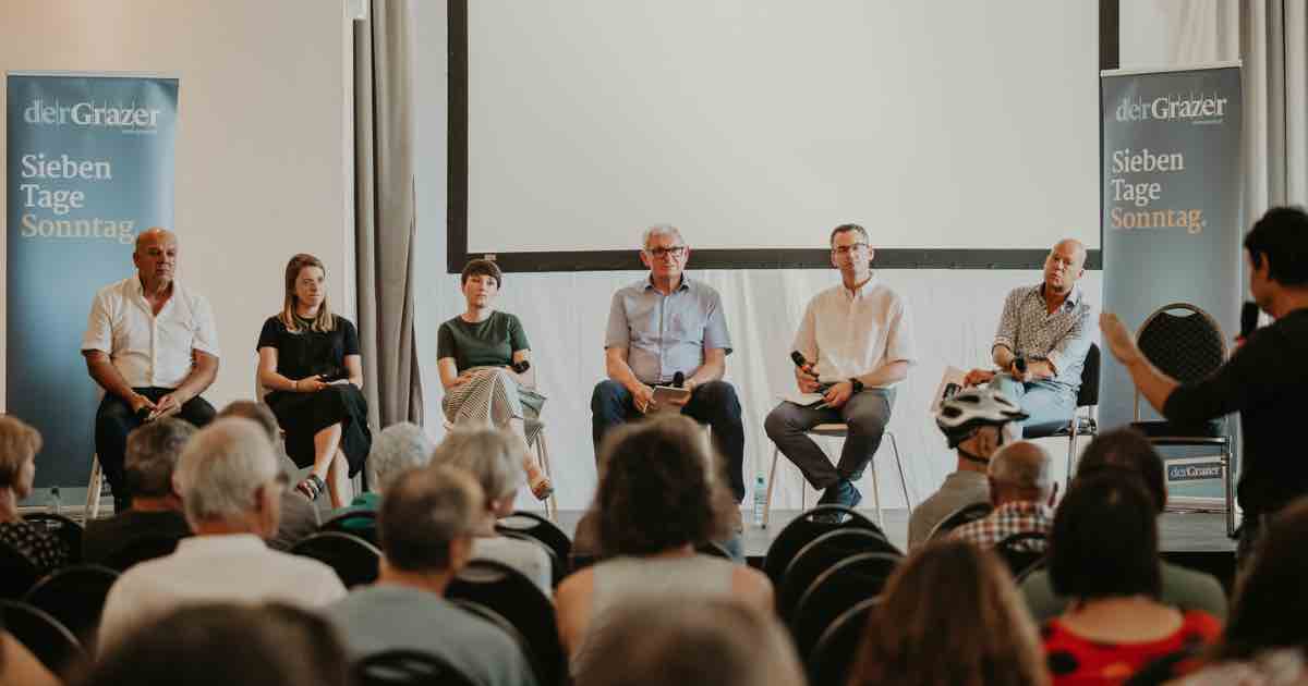 Sechs Panelteilnehmende sitzen auf einer Bühne bei einer Diskussionsrunde im Saal, vor Publikum; Banner links und rechts, Leinwand im Hintergrund.