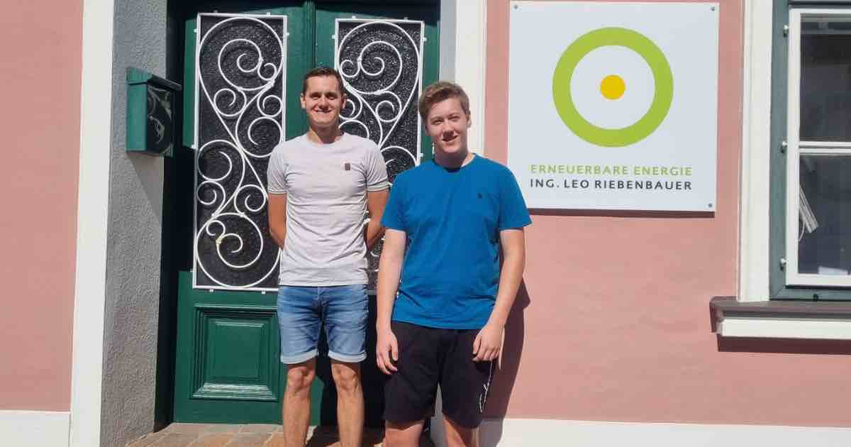 Zwei Personen stehen vor einer Tür mit schmiedeeisernen Mustern; rechts Schild mit grünem Ringlogo und Text 'Erneuerbare Energie'.