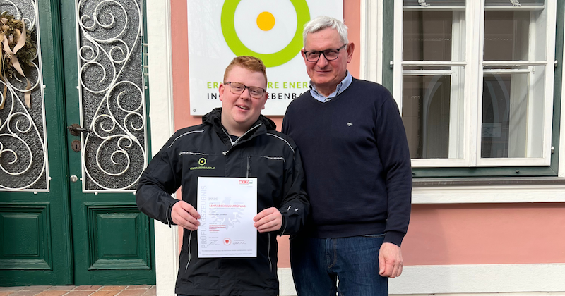 Zwei Personen stehen vor einem Gebäude; eine Person hält eine Urkunde, die andere Person steht daneben.