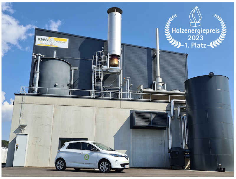 Industrieanlage zur Holzenergie mit Tanks, Rohrleitungen und Fassade; weißes Auto davor; Logo Holzenergiepreis 2023 1. Platz oben rechts.