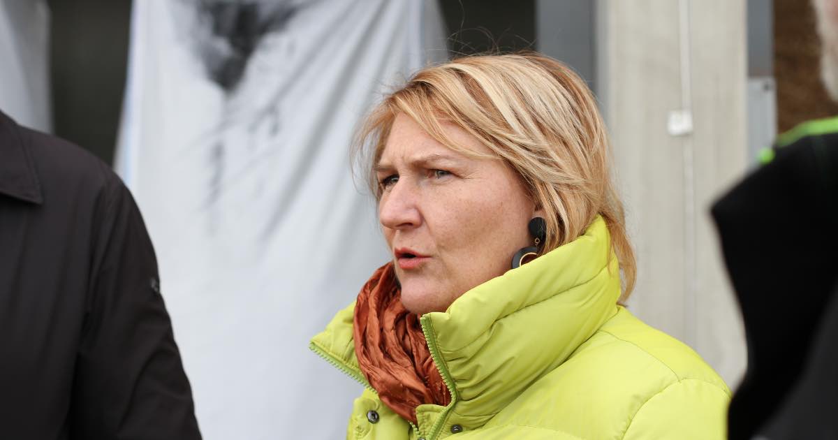 Person mit blonden Haaren, limegrüne Jacke und orangefarbenem Schal, trägt schwarzen Ohrring, spricht im Außenbereich.