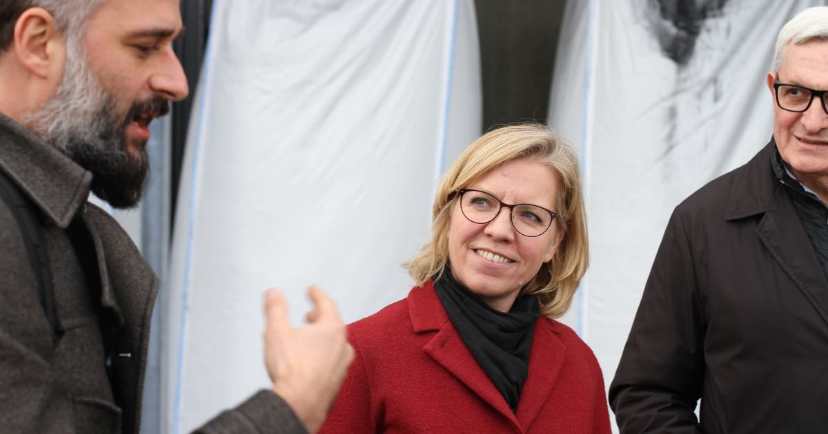 Drei Personen im Gespräch: links eine Person mit Bart, mitte eine Person mit Brille in rotem Mantel, lächelnd, rechts eine Person in dunkler Jacke.