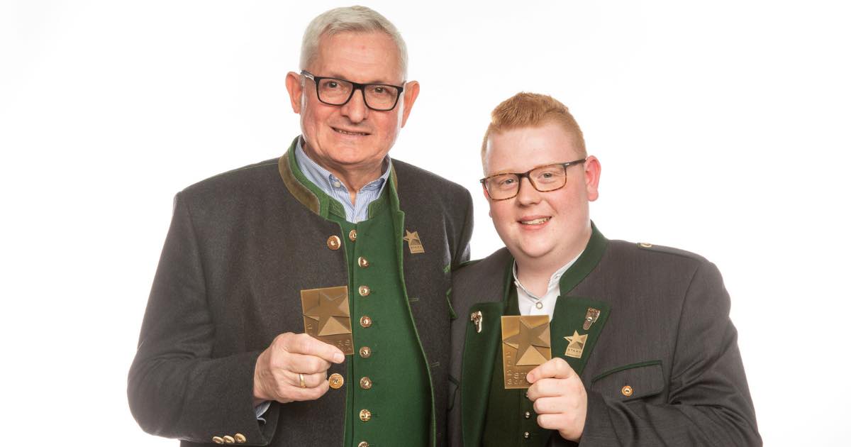 Zwei Personen in traditioneller Trachtenkleidung halten goldene Abzeichen mit Sternmotiv.