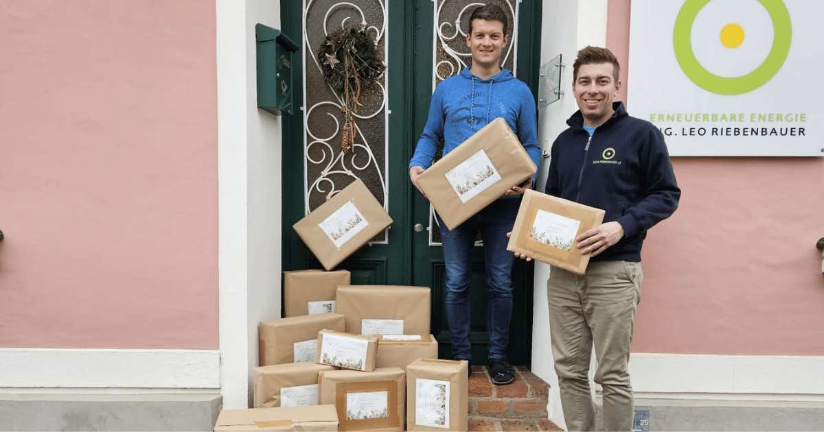 Zwei Personen stehen vor einer Tür und halten braune Kartonpakete; davor liegen weitere verpackte Sendungen auf Stufen.