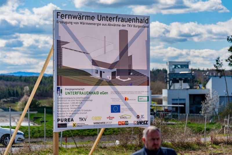 Informationsschild Fernwärme Unterfrauenhaid zeigt Gebäude-Silhouette und Kamin; Outdoor-Bauprojekt, unscharfe Person im Vordergrund.