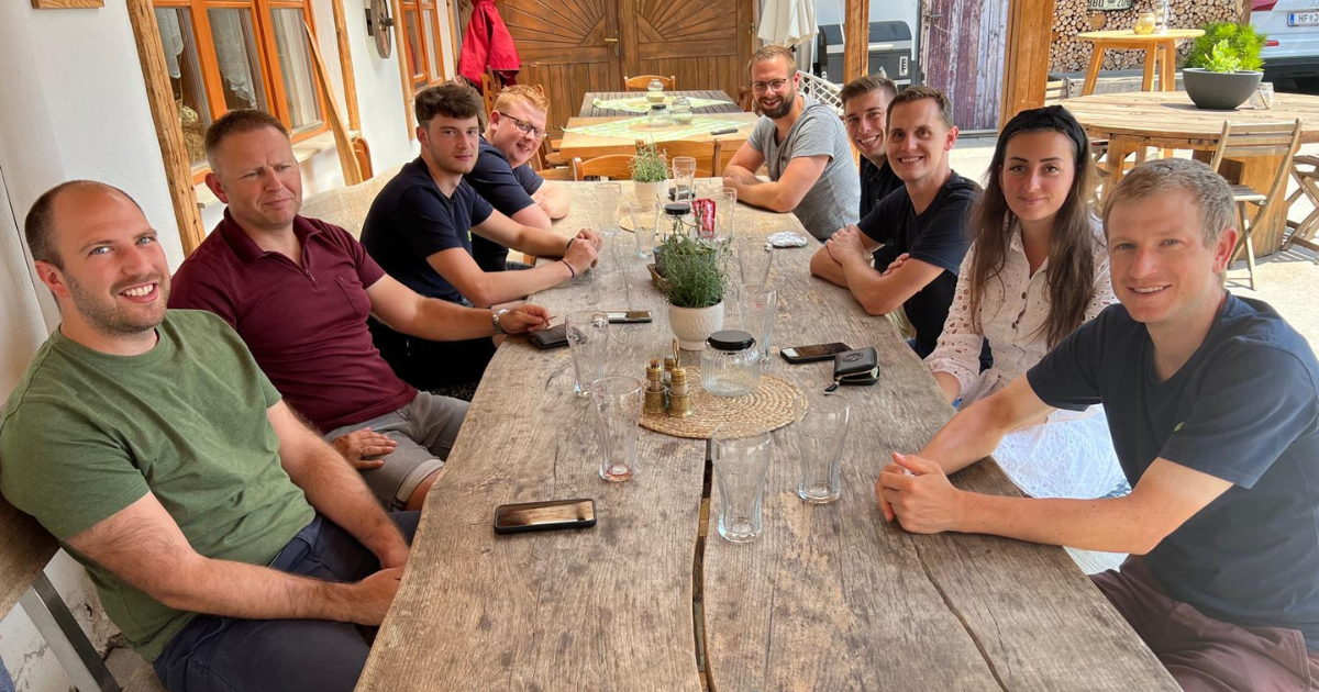 Gruppe von Personen sitzt an einem langen Holztisch im Freien, lächelt, mit Gläsern und Pflanzendekoration.