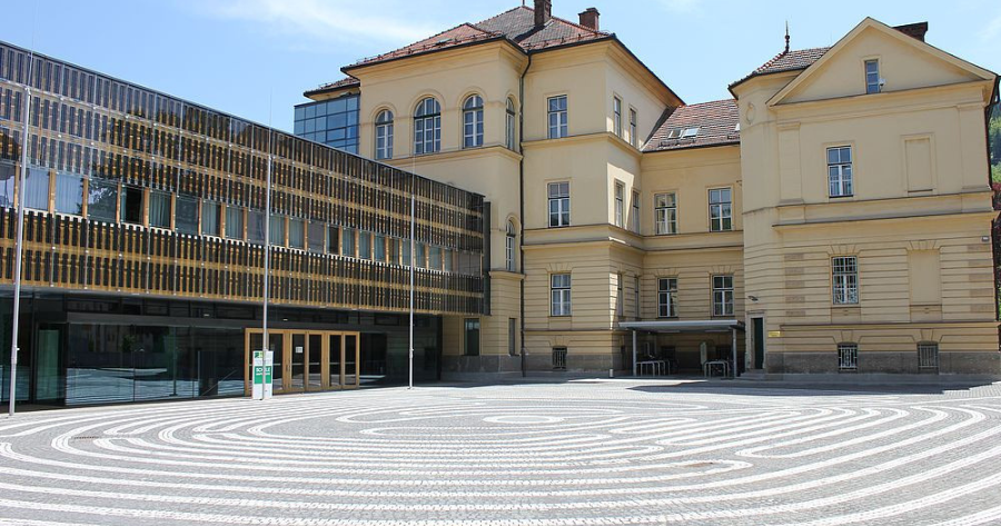 Historische gelbe Gebäudefassade neben moderner Glasfassade; gepflasterter Platz mit konzentrischen Kreismustern vor dem Eingang.
