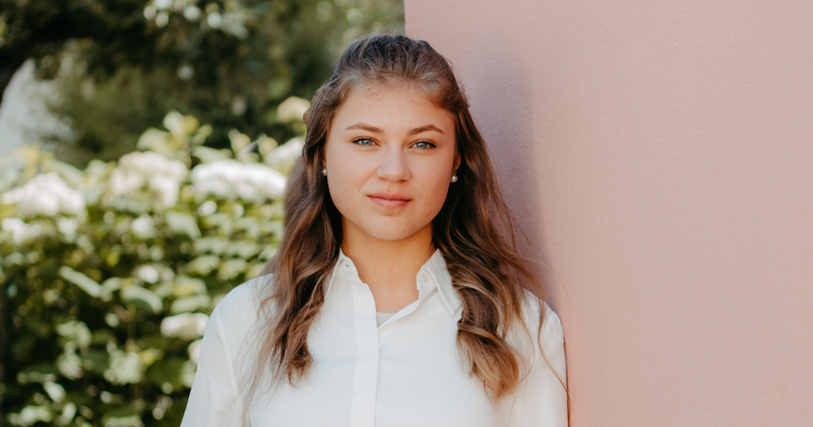 Person mit blonden Haaren und weißem Hemd steht an rosa Wand, grüne Gartenlandschaft im Hintergrund.