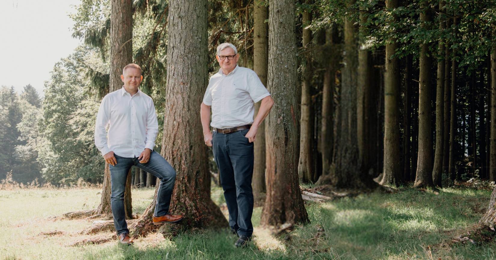 Zwei Personen in weißen Hemden stehen zwischen Baumstämmen in einem Wald; eine lehnt an einem Stamm, die andere hat die Hände in den Taschen.