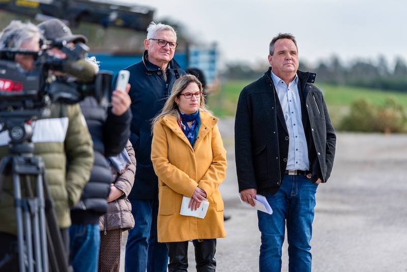 Pressegruppe im Freien mit Kamerateam; drei zentrale Personen stehen vorne, eine im senfgelben Mantel, eine in dunkler Jacke, hält Notizen.