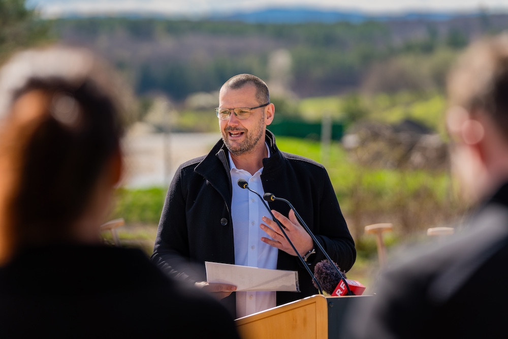 Person spricht hinter Rednerpult im Freien, hält Unterlagen, trägt Brille und dunkle Jacke; Mikrofone vor Ort, unscharfe Personen im Vordergrund.