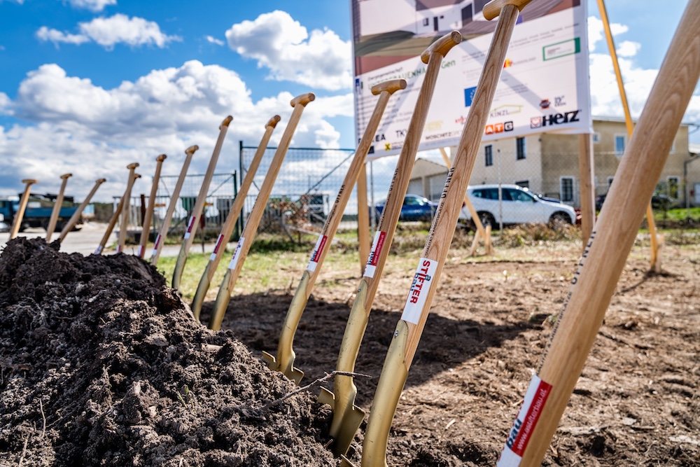 Mehrere Spaten mit Holzstielen stehen in gegrabener Erde auf einer Baustelle; Hintergrund mit Schild, Zaun und Fahrzeugen.