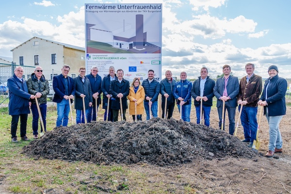Gruppe von Personen beim Spatenstich für ein Fernwärmeprojekt, Erdhaufen im Vordergrund, Banner mit Projektdetails im Hintergrund.