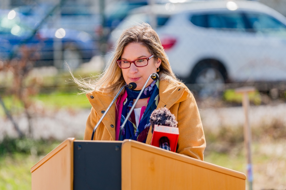 Person hinter Rednerpult im Freien, gelbe Jacke, bunter Schal, rote Brille, spricht in Mikrofone bei einer Pressekonferenz.