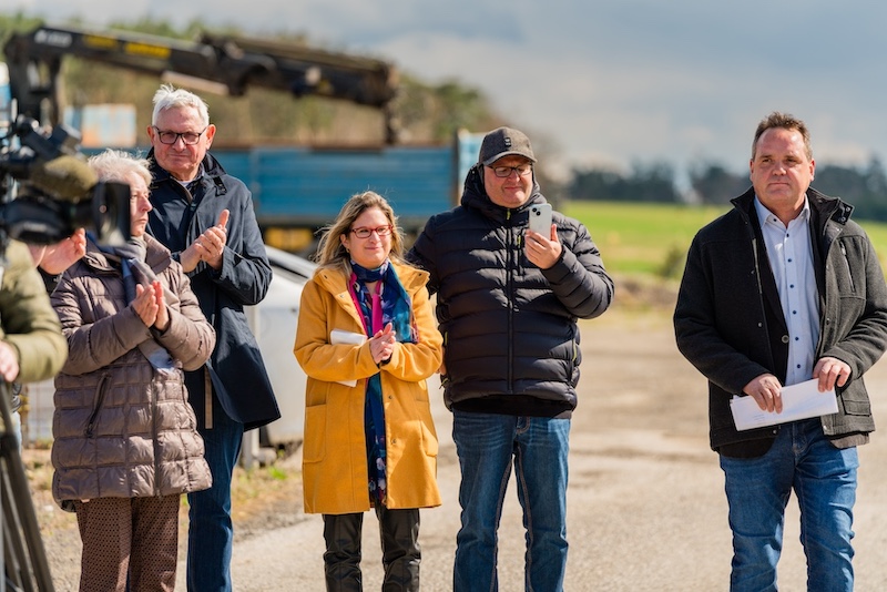 Gruppe von sechs Personen draußen; eine Person hält Smartphone, eine weitere trägt gelben Mantel; andere klatschen, Ausrüstung sichtbar im Hintergrund.