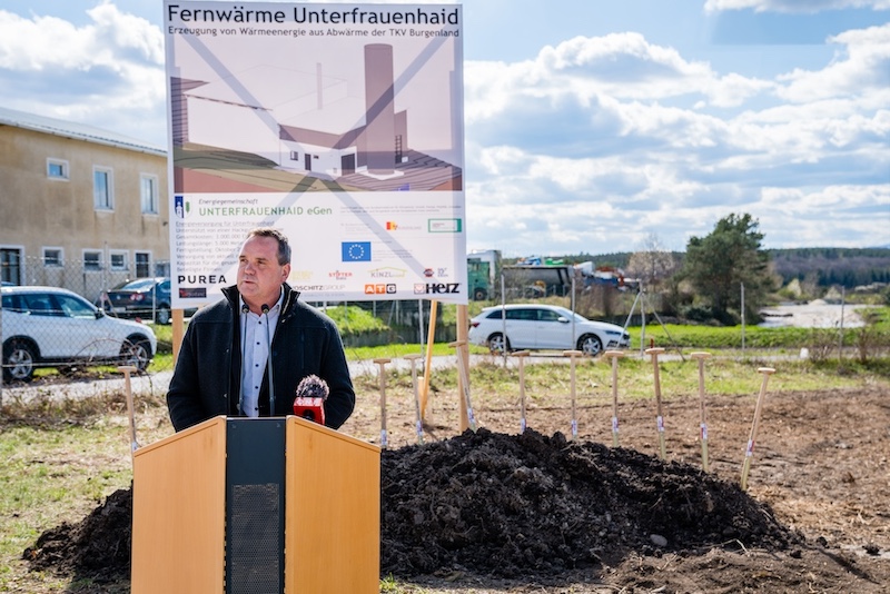 Eine Person spricht hinter einem Rednerpult auf einer Baustelle; im Hintergrund ein Plakat zur Fernwärme Unterfrauenhaid und Erdarbeiten.