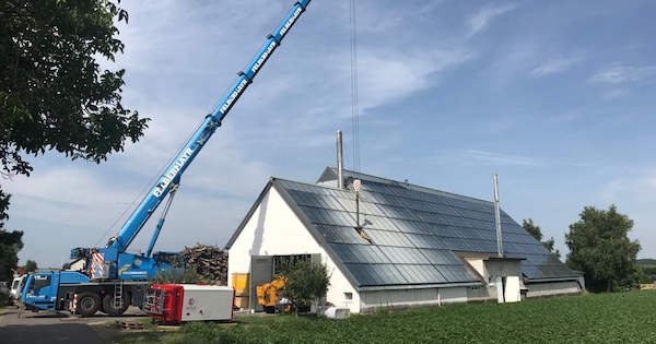 Blauer Kran hebt Material neben einem Gebäude mit schrägem Dach auf einer Baustelle; Holzstapel und Baugeräte im Grünland.