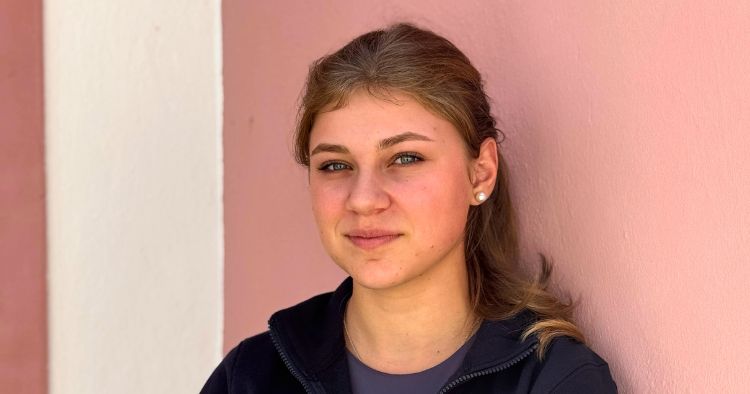 Portrait einer Person vor einer rosa Wand, braune Haare, dunkle Jacke, neutraler Blick in die Kamera.