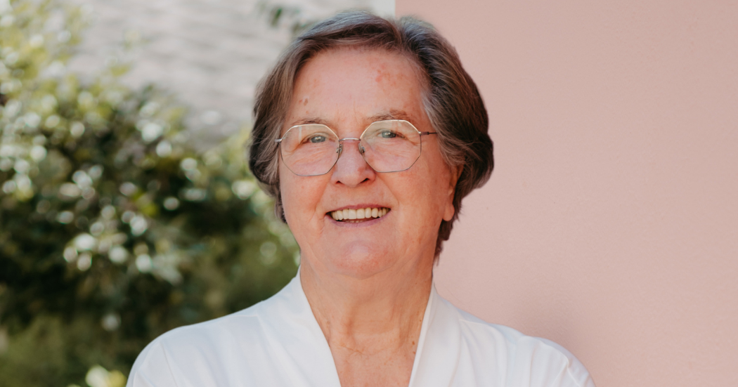 Person mit kurzen grauen Haaren, Brille und weißem Oberteil, lächelnd vor unscharfem Hintergrund aus Grünpflanzen und rosa Wand.