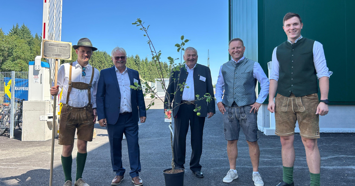 Fünf Personen stehen draußen in Trachten- und Businesskleidung um einen kleinen Baum im Topf.