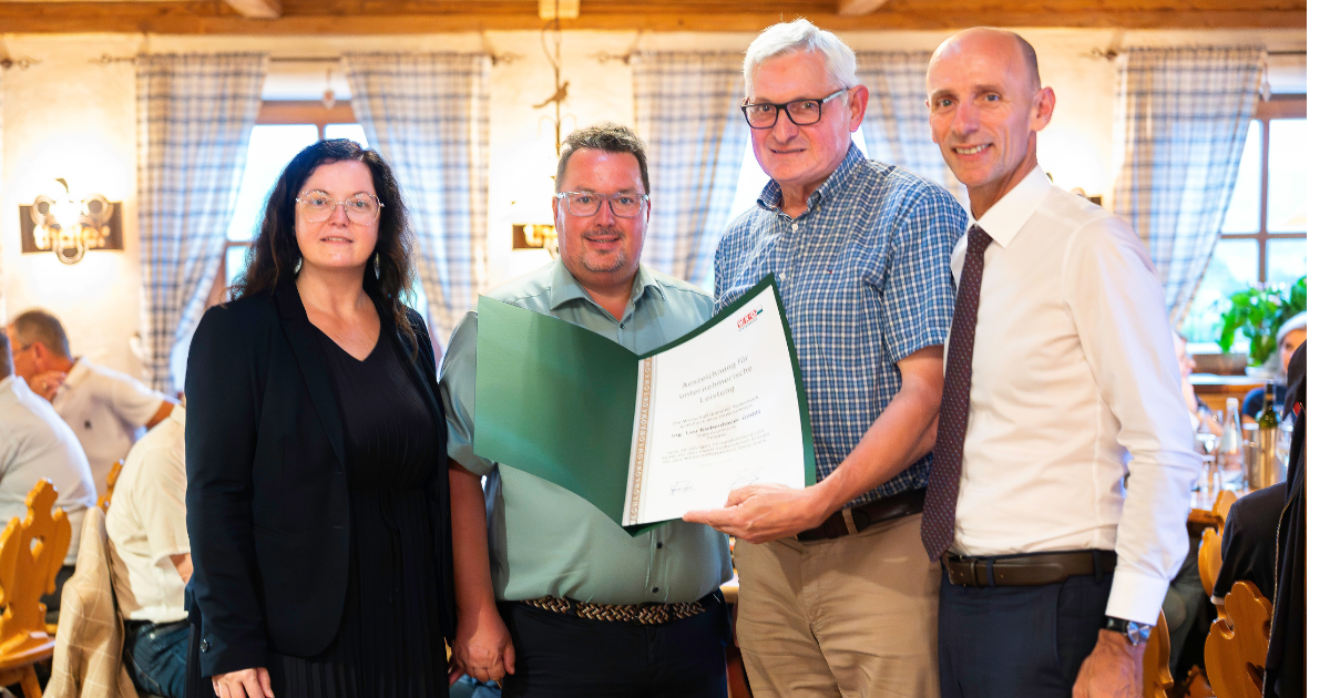 Vier Personen stehen zusammen; eine Person präsentiert eine Urkunde in einem Ordner bei einer formellen Veranstaltung im Innenraum.