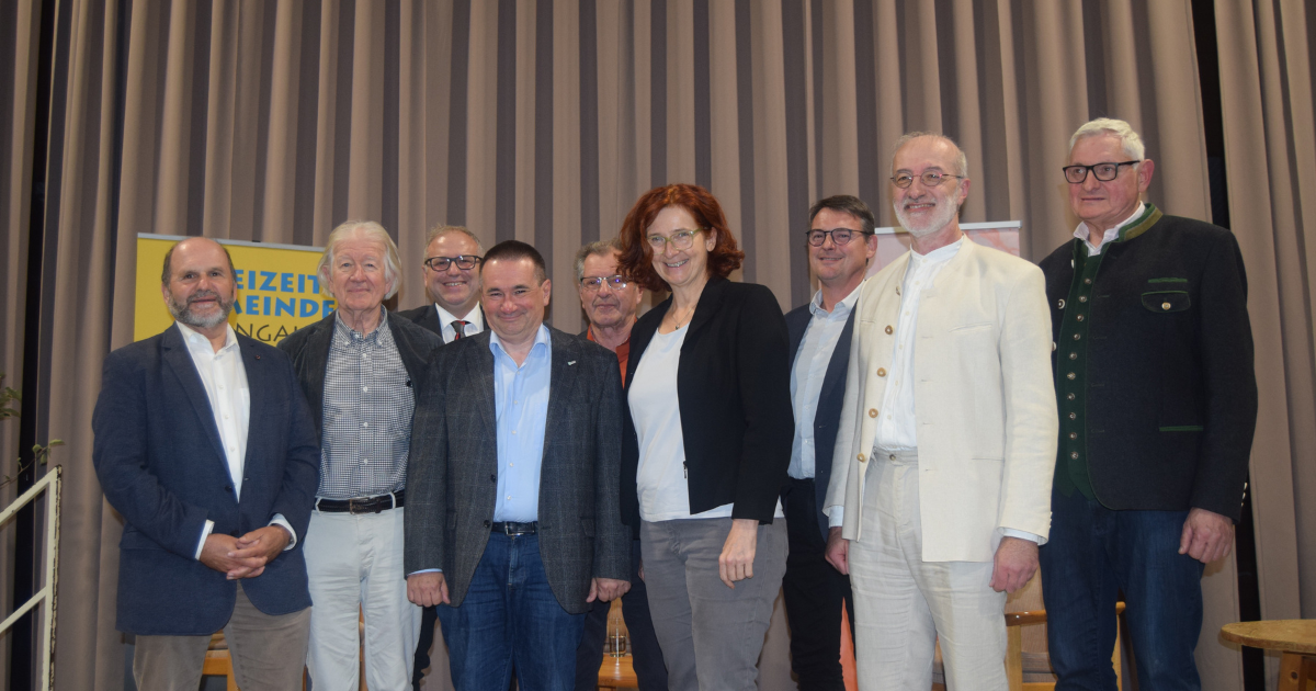 Gruppe von Personen posiert auf einer Bühne vor grauem Vorhang; unterschiedliche formelle Outfits, freundlich in die Kamera blickend.