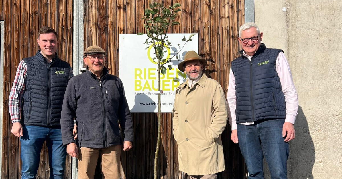 Vier erwachsene Personen stehen nebeneinander vor einer Holzwand mit Schild und gelber Schrift; davor eine Pflanze, alle schauen in die Kamera.