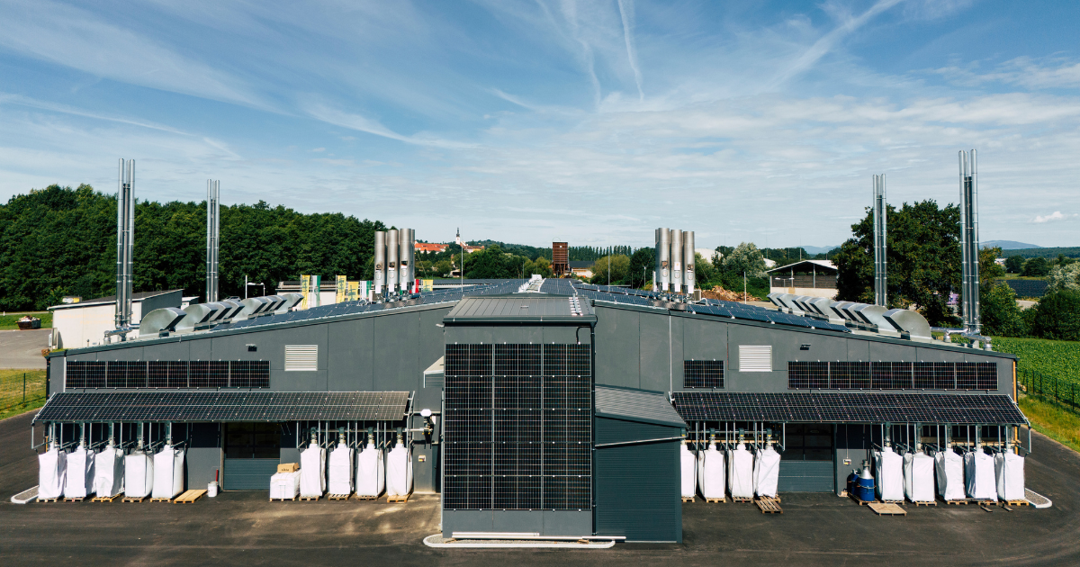 Großes Industriegebäude mit PV-Modulen an Fassaden, Silos, hohen Schornsteinen und Paletten mit weißen Säcken, umgeben von Feldern und Bäumen.