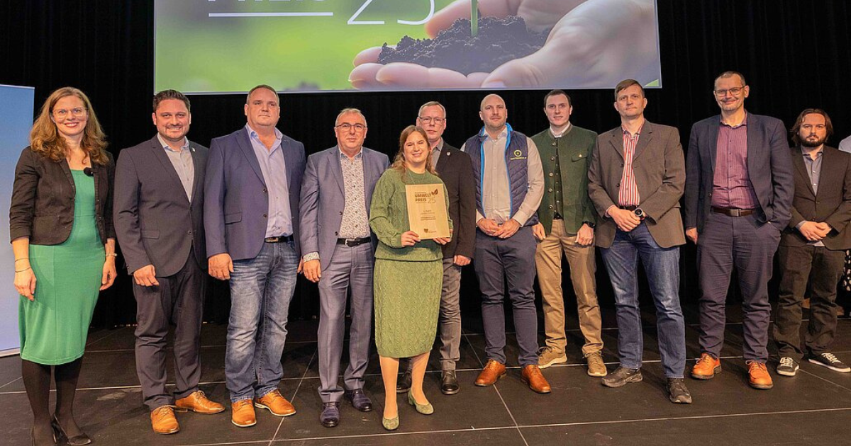 Gruppe von Personen steht auf einer Bühne; zentrale Person hält Urkunde bei Preisverleihung.