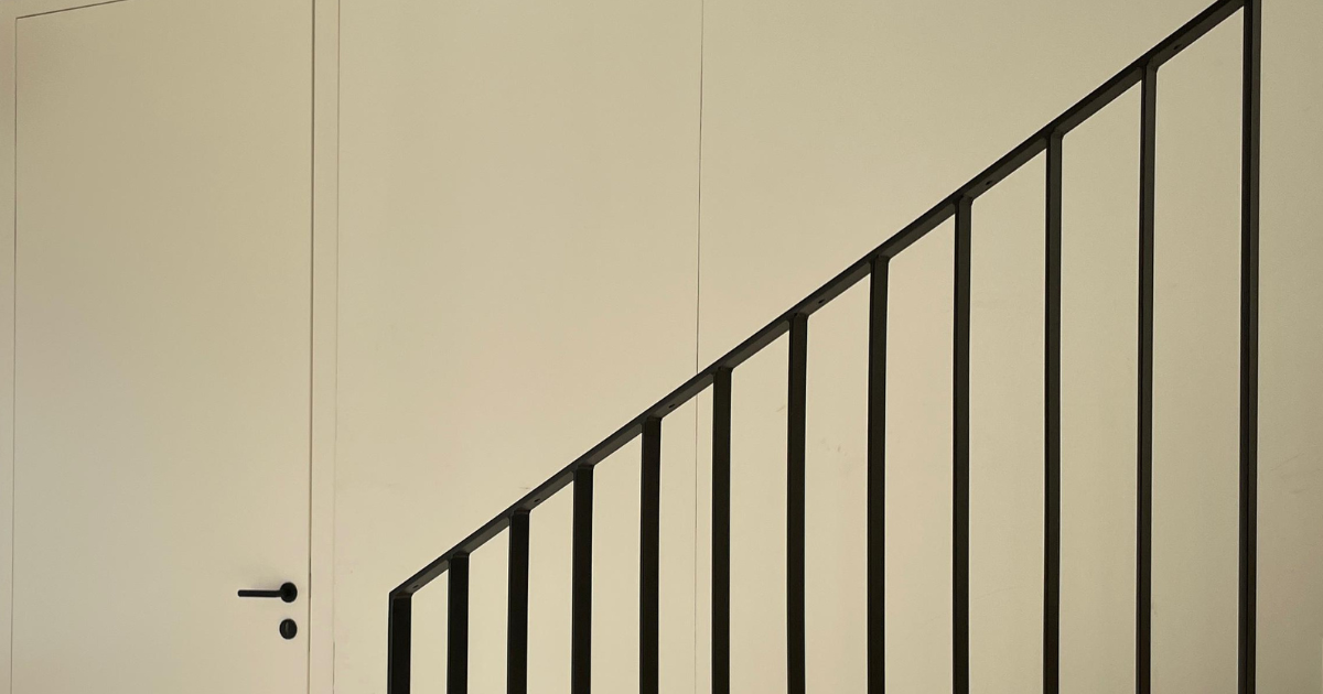 Weiße Innenwand mit schwarzem Metallgeländer einer diagonal verlaufenden Treppe; linke Tür mit Griff sichtbar.