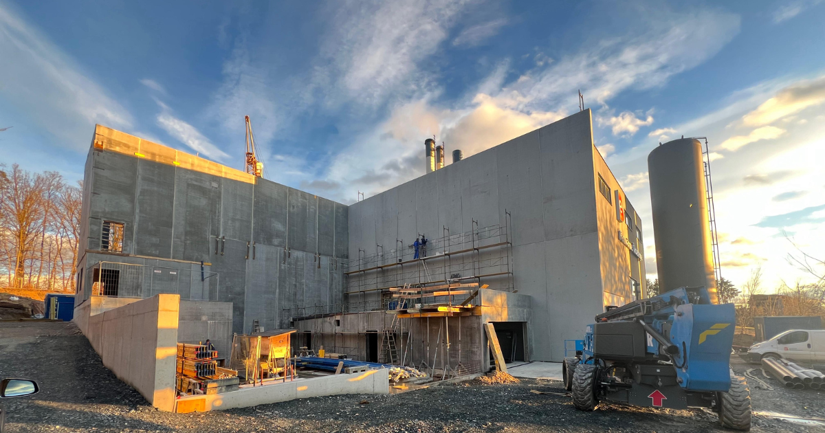 Große Betonbaustelle mit Gerüsten und Baugeräten, eine Person auf dem Gerüst; Kran im Hintergrund, sonniger Himmel.