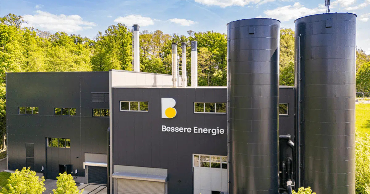 Moderne Industrieanlage mit zwei großen schwarzen Pufferspeichern, Gebäude mit dem Logo der Burgenland Energie und grünem Umfeld.