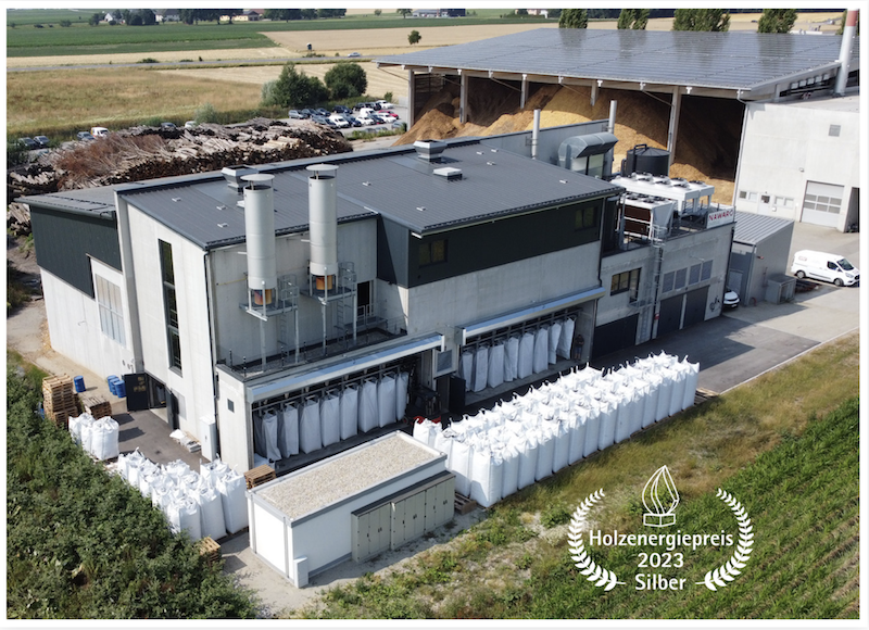 Außenansicht einer Holzenergieanlage mit Pufferspeichern, Sacklagern und dem Siegel Holzenergiepreis 2023 Silber.