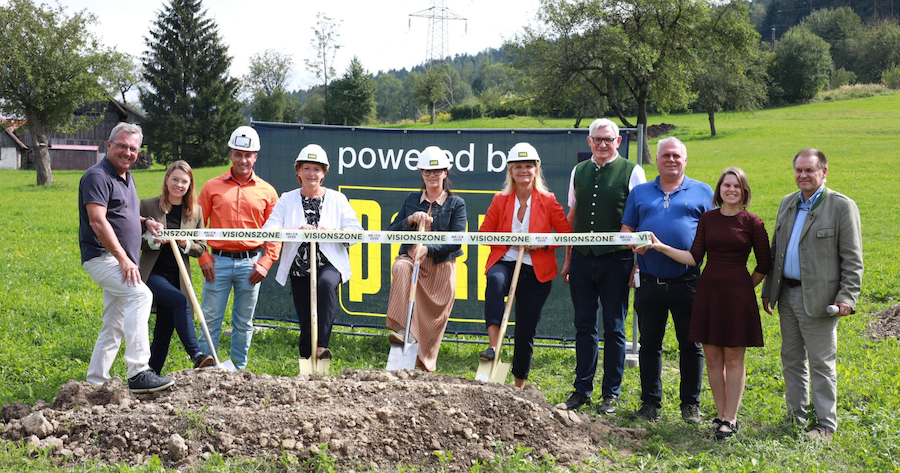 Gruppe beim Spatenstich auf grünem Gelände, Spaten, Absperrband 'VISIONZONE' und Banner 'powered by' im Hintergrund.